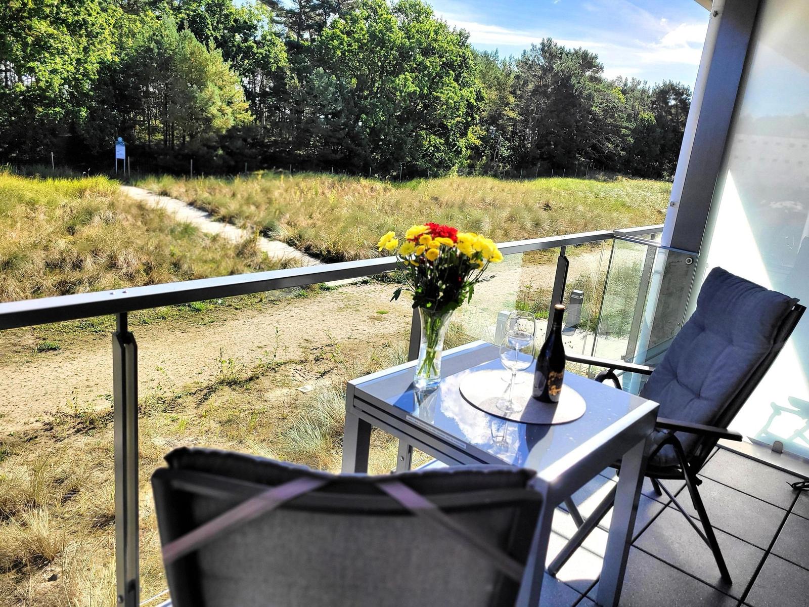 Terrasse mit Tisch, Stühlen, Blumen und Flasche. Blick auf Grasland und Wald.