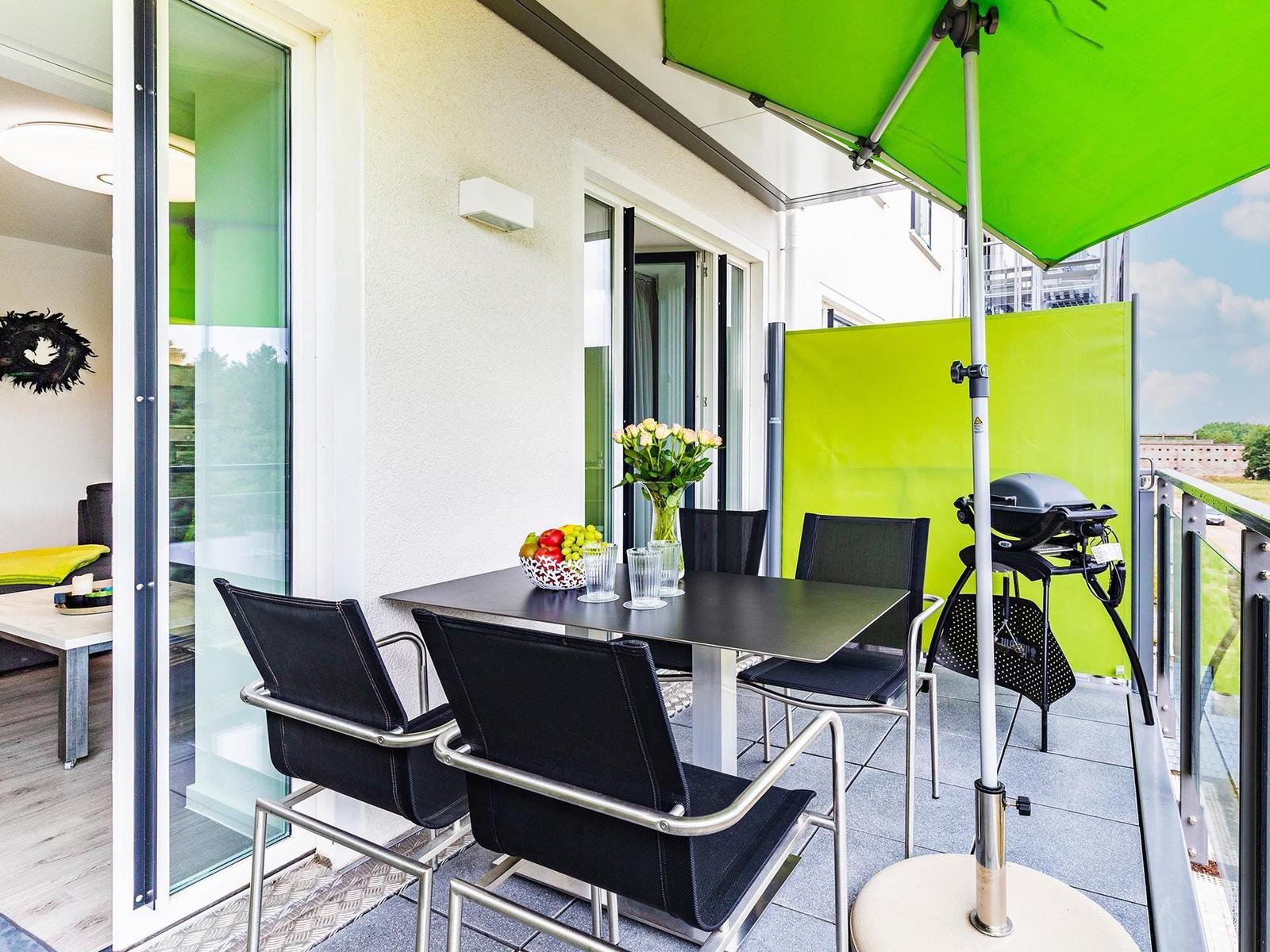 Terrasse mit Tisch, Stühlen, Grill und grünem Sonnenschirm. Blick auf grüne Landschaft.