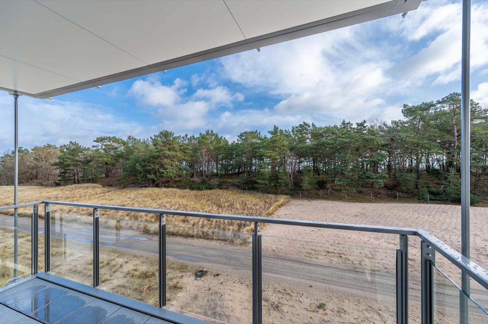 Terrace with glass railing offers view of sandy beach and forest.