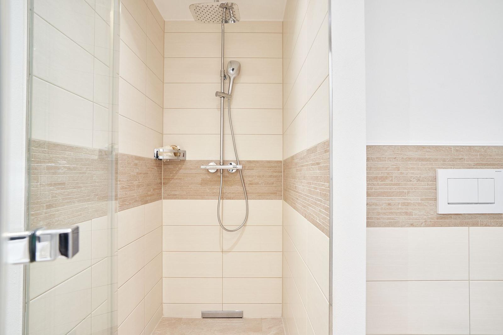 Duschkabine mit Glaswand, Duschbrause und Handbrause. Wandfliesen in Beige und Braun.