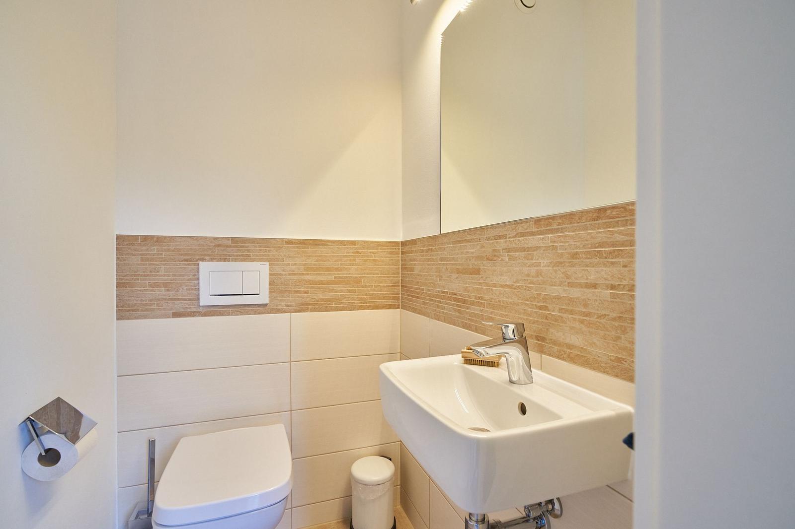Badezimmer mit Toilette, Waschbecken und Spiegel. Wandverkleidung aus hellen Fliesen.