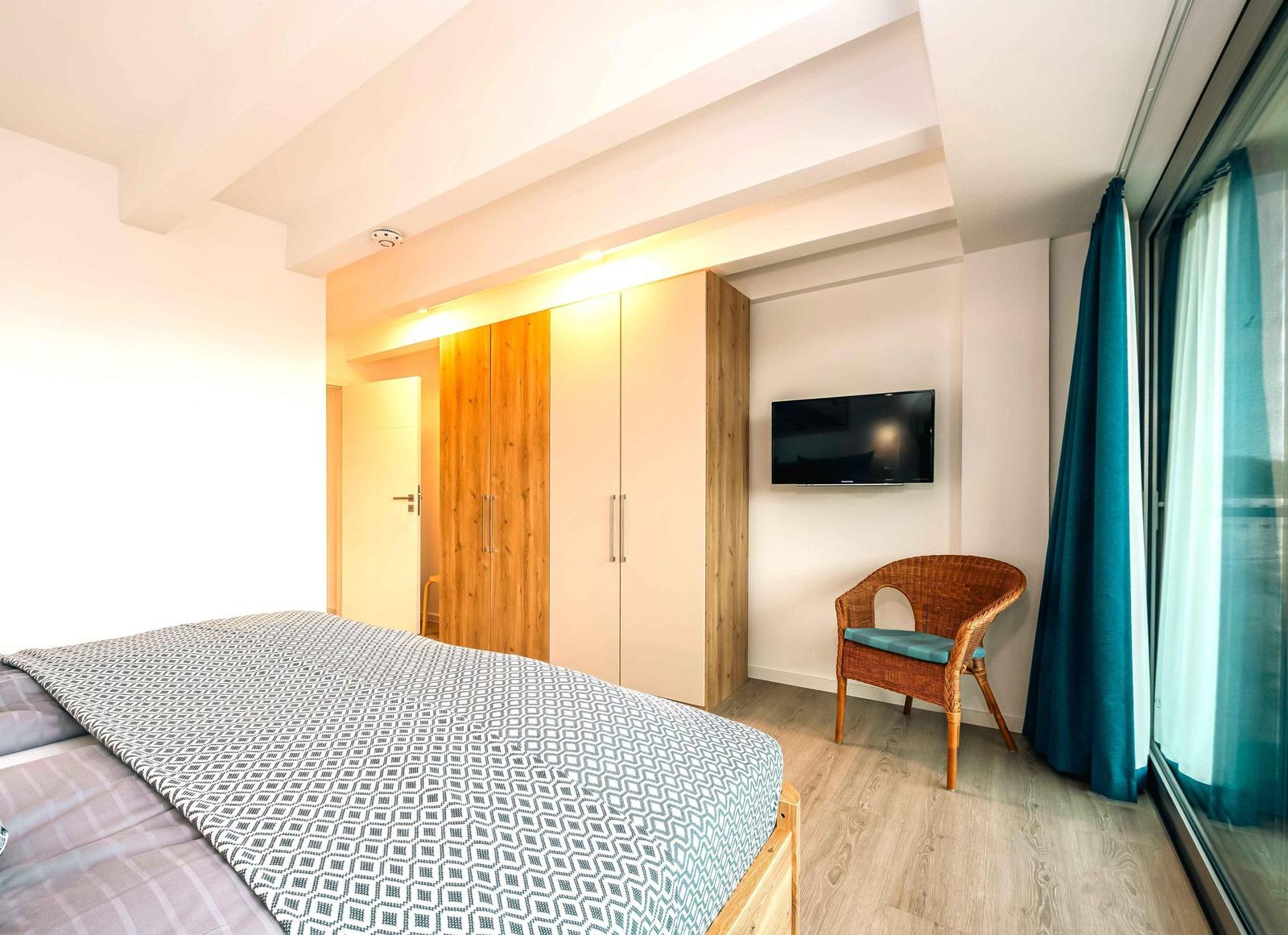 Schlafzimmer mit Bett, Kleiderschrank, TV und Sessel. Große Fenster mit Vorhängen.