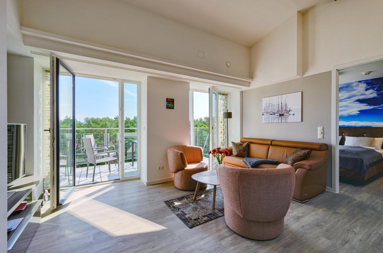 Living room with sofa, chairs, and balcony. Bedroom with bed in background.