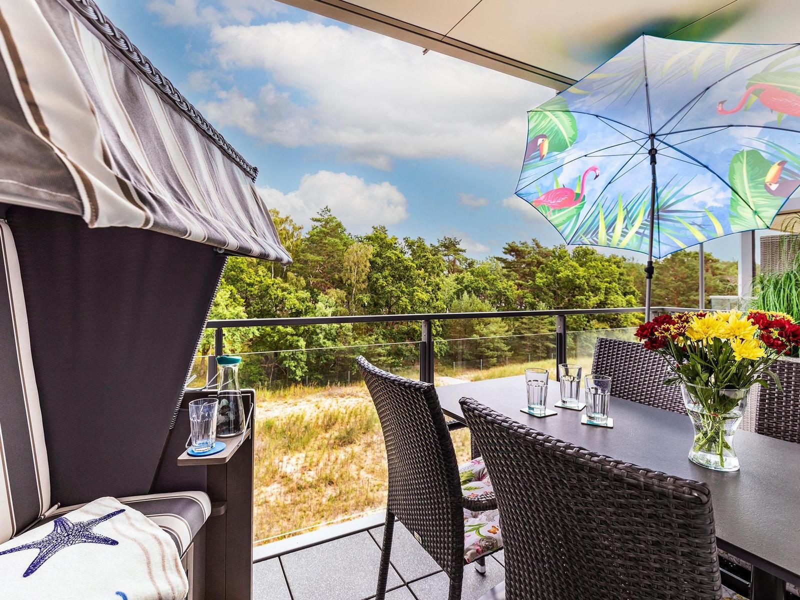 Terrasse mit Sitzgruppe, Sonnenschutz und Tisch mit Blumen. Blick auf grüne Landschaft.