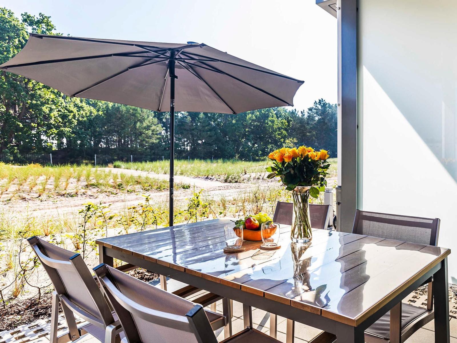 Terrasse mit Tisch, Stühlen und Sonnenschirm. Blumen und Obst auf dem Tisch.