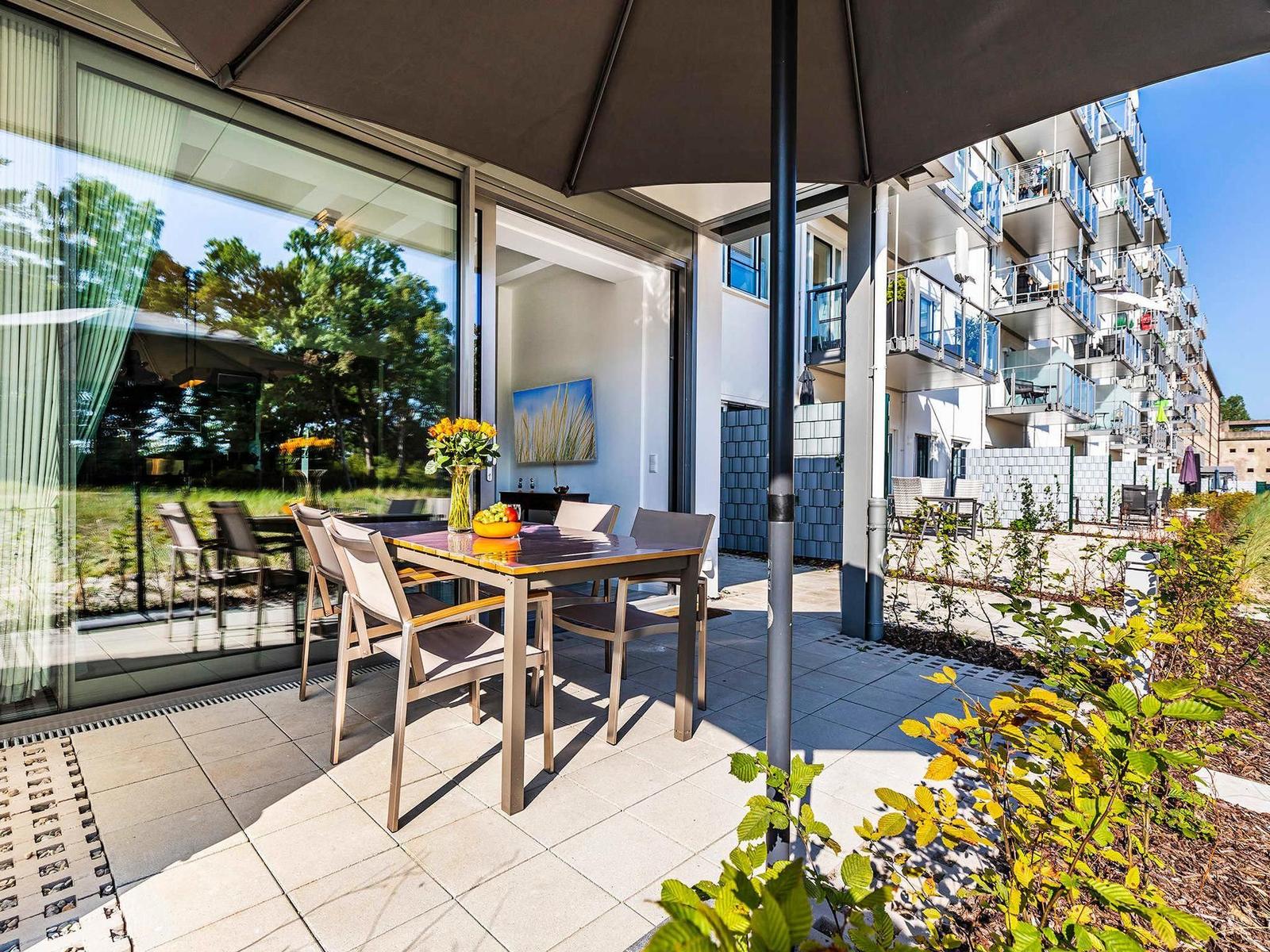 Terrasse mit Tisch, Stühlen und Sonnenschirm. Glasfront führt ins Haus. Hinterhof mit Wohngebäude.