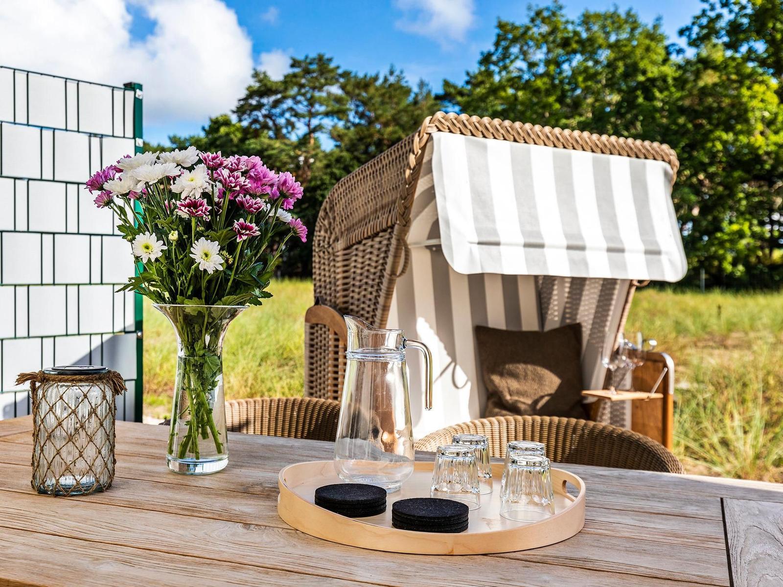 Holztisch mit Blumen, Wasserglas und Strandkorb im Hintergrund.