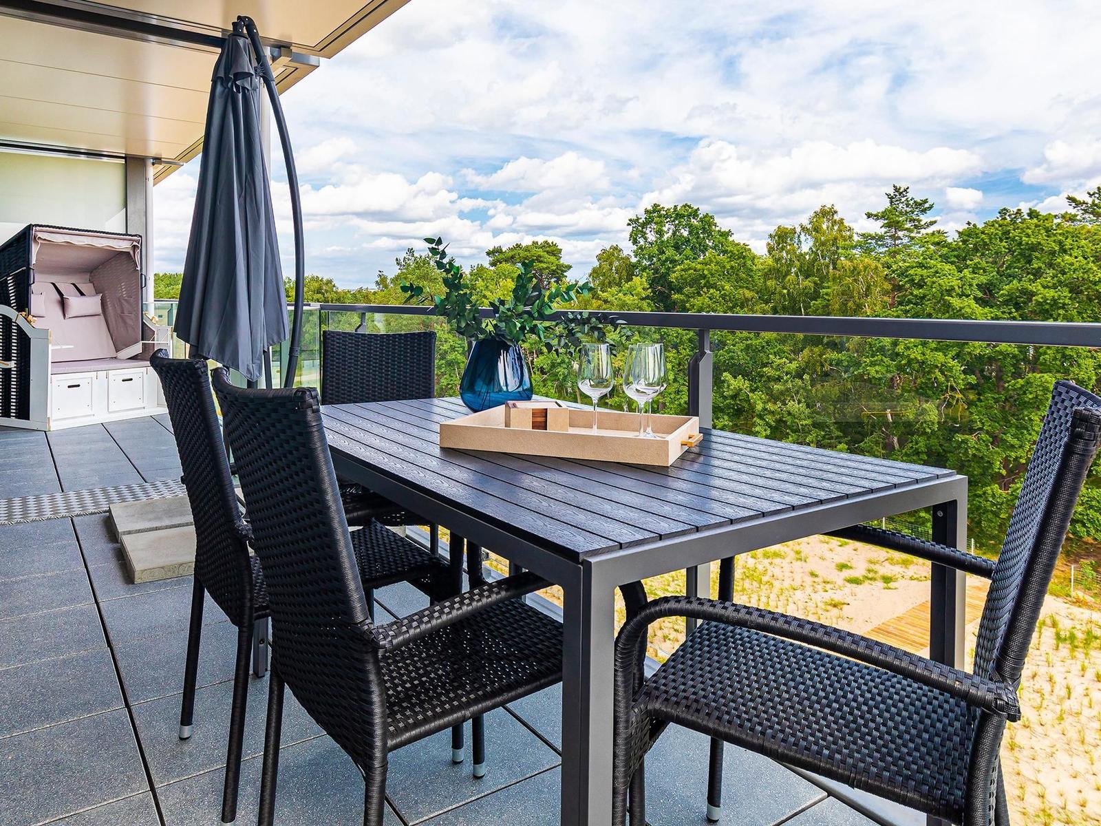 Terrasse mit Tisch, Stühlen und Sonnenschirm. Blick auf grüne Bäume und Himmel.