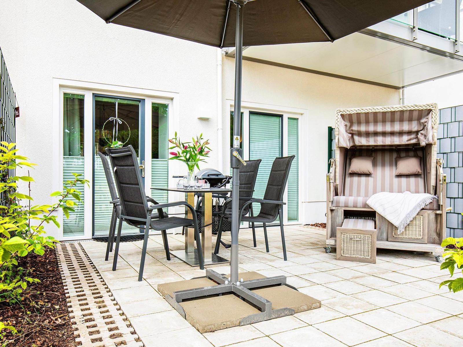 Terrace with table, chairs, umbrella, and beach chair. Background: house with windows.