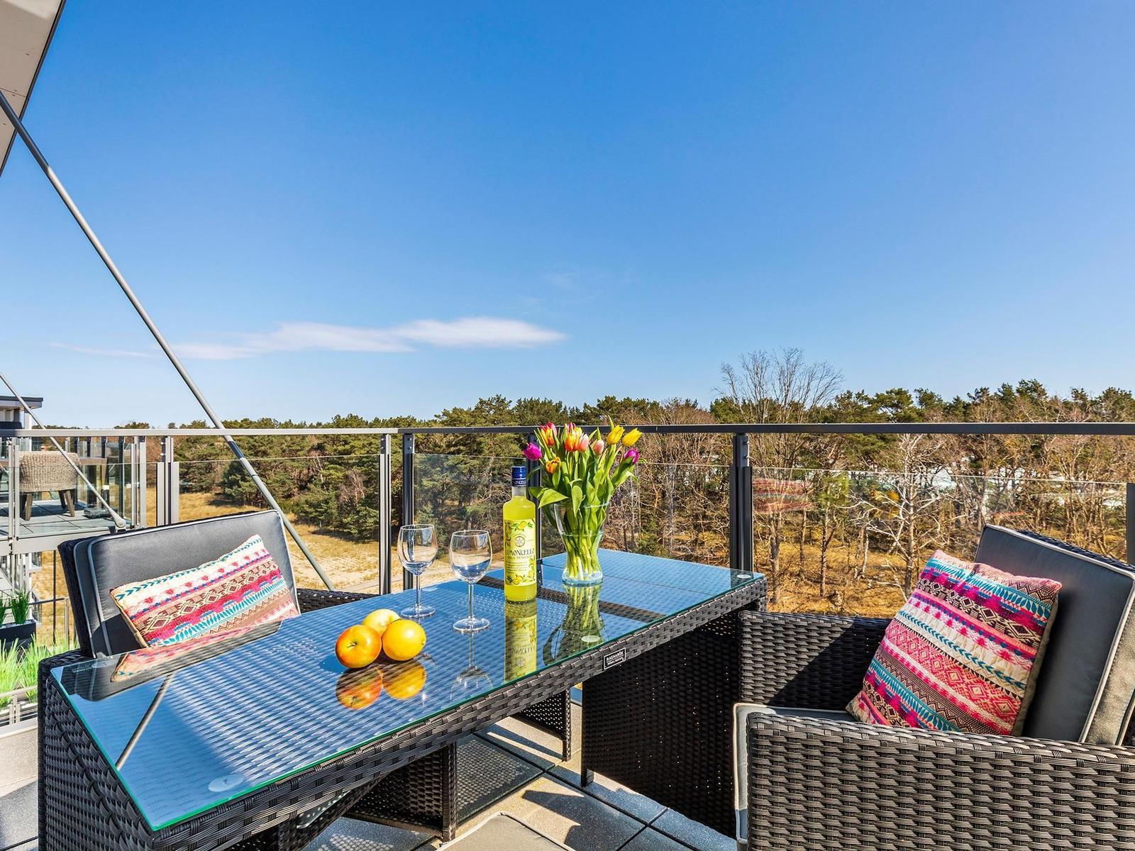 Terrasse mit Tisch, Stühlen und Blumen. Blick auf Wald und Himmel.