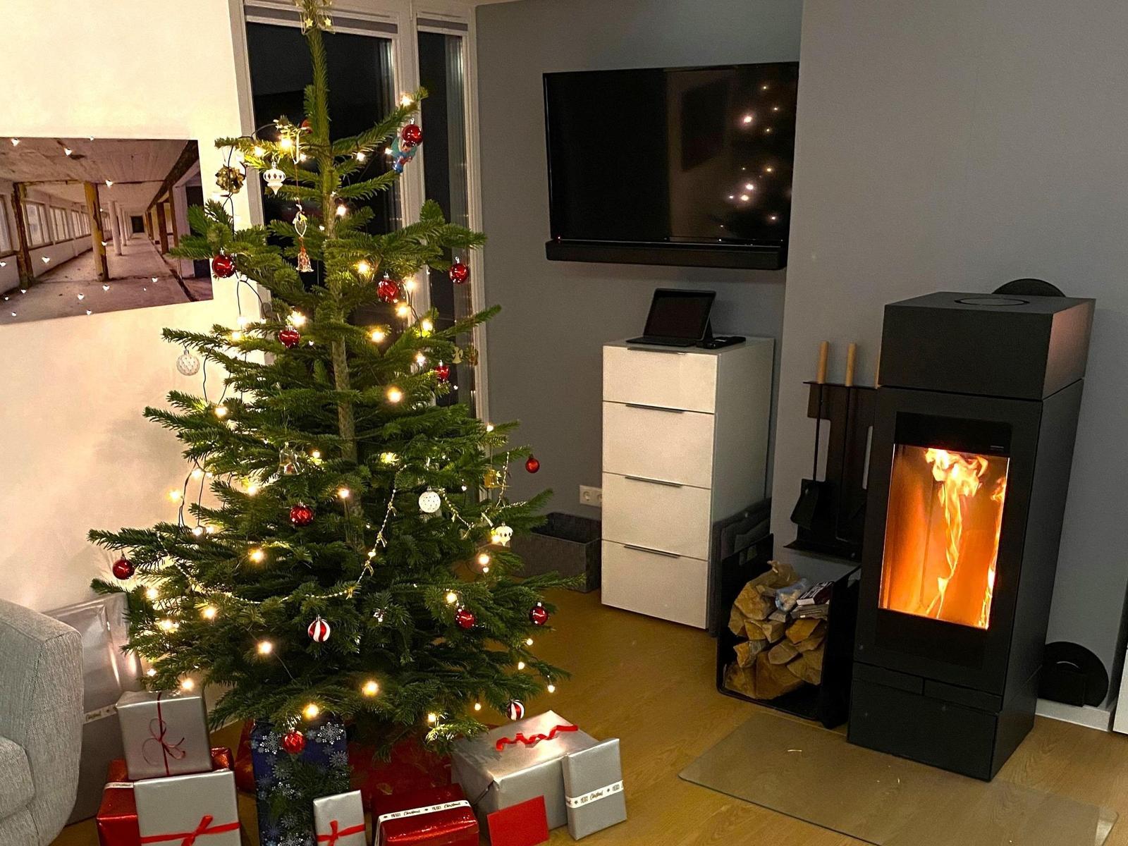 Weihnachtsbaum mit Lichtern und Geschenken neben Kamin und Fernseher.