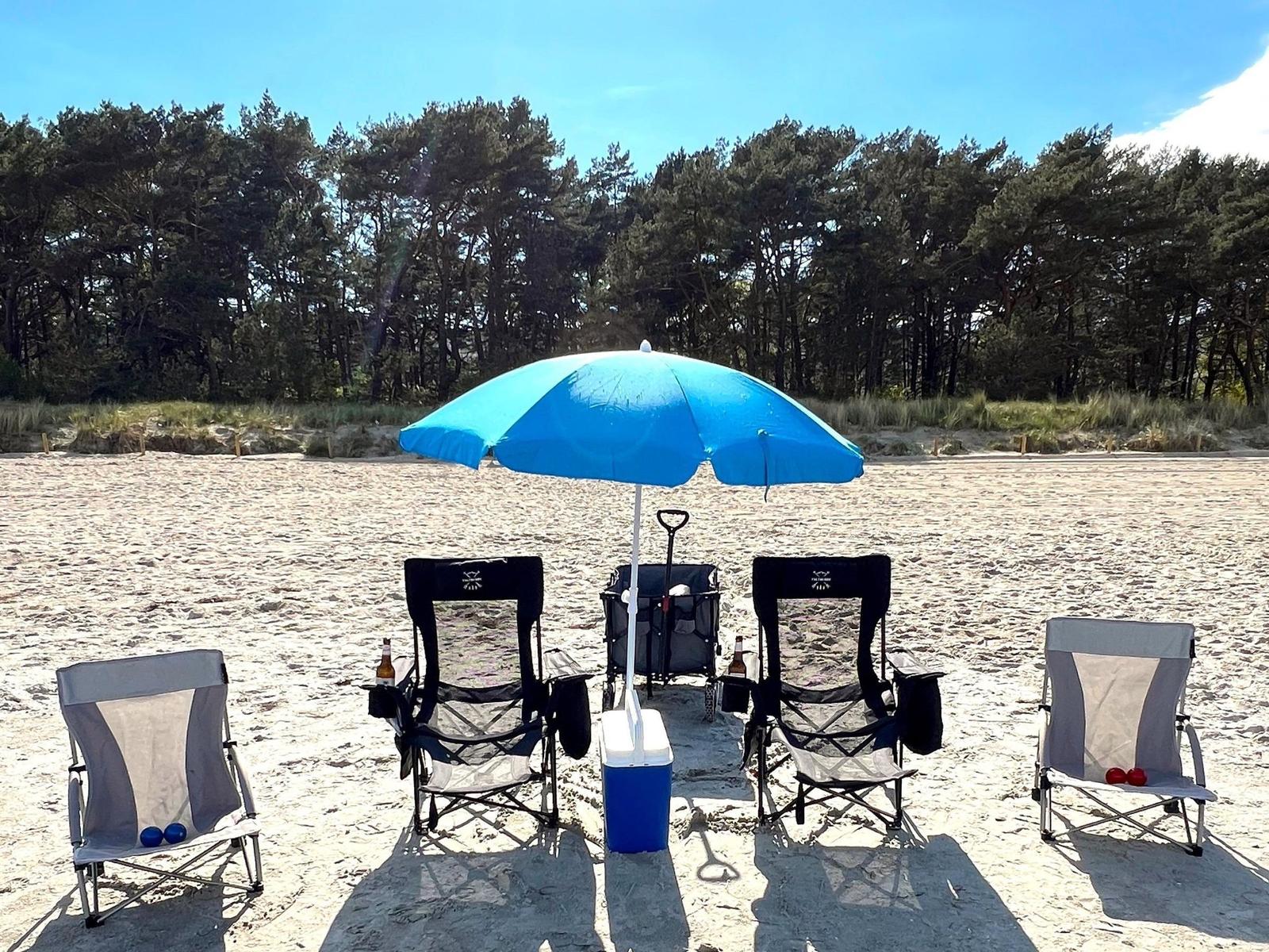 Blauer Strandparasol mit Stühlen und Kühler auf Sandstrand vor Wald.