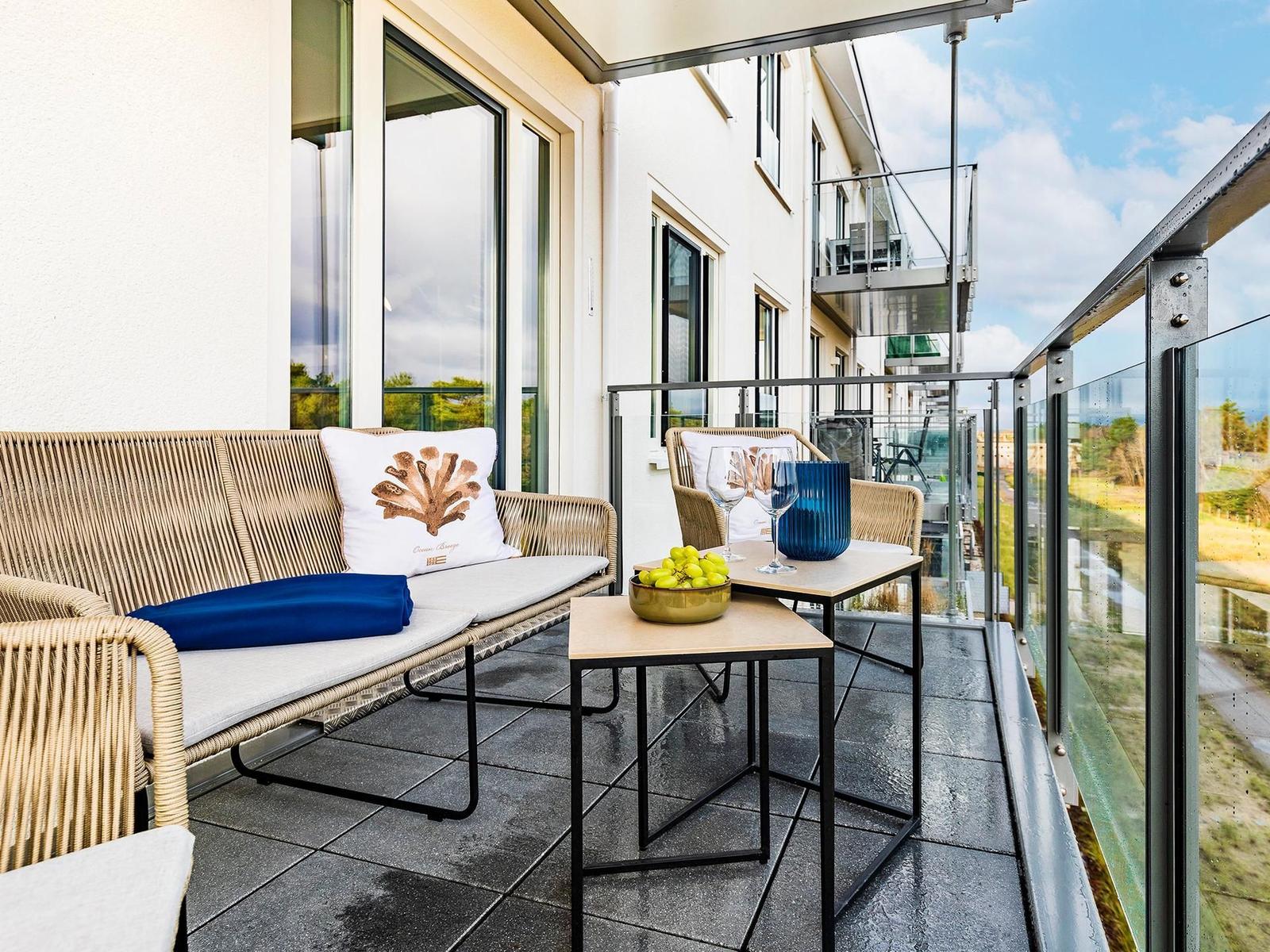 Terrasse mit Rattanmöbeln, Tisch, Gläsern und Obst. Glasgeländer und Blick auf Gebäude.
