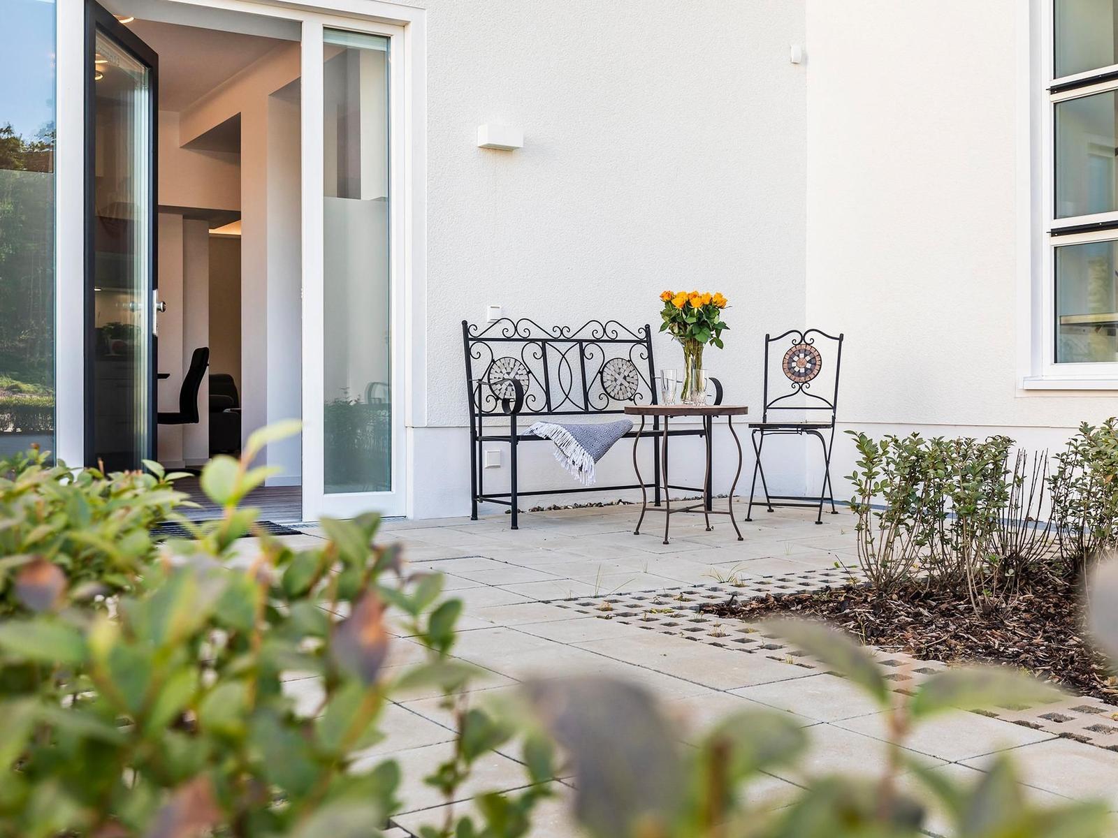 Terrasse mit Tisch, Bank und Stuhl. Blumen und Pflanzen umgeben den Außenbereich.
