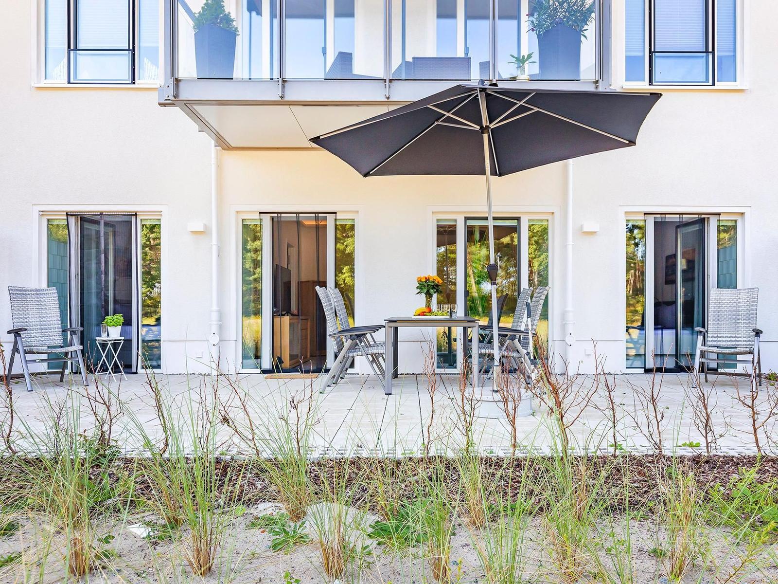 Terrasse mit Tisch, Stühlen und Sonnenschirm. Glasfronten führen ins Innere. Vordergrund: Gras und Sand.