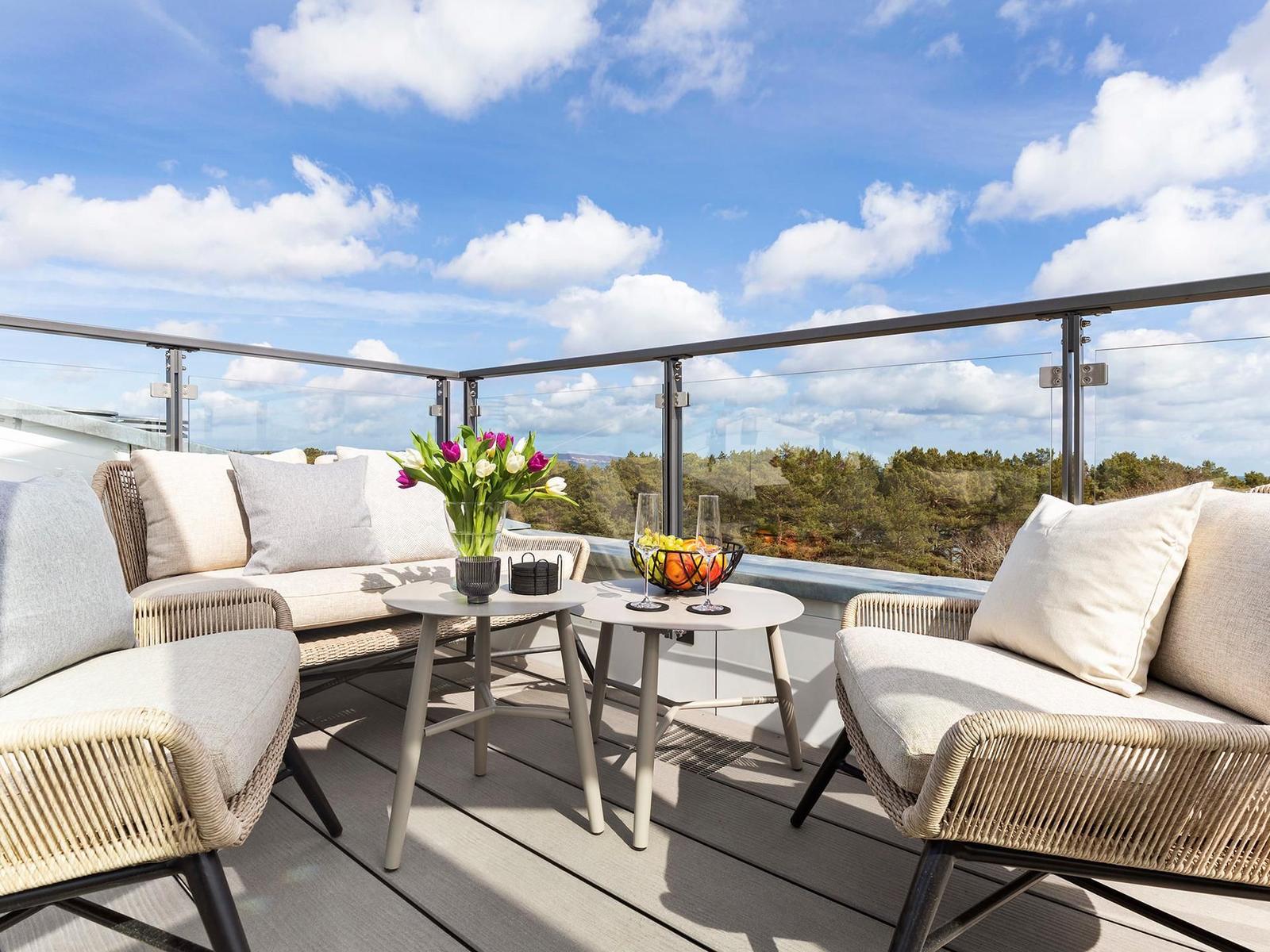 Terrasse mit Sitzgruppe, Tisch und Blumen. Blick auf Wald unter blauem Himmel.