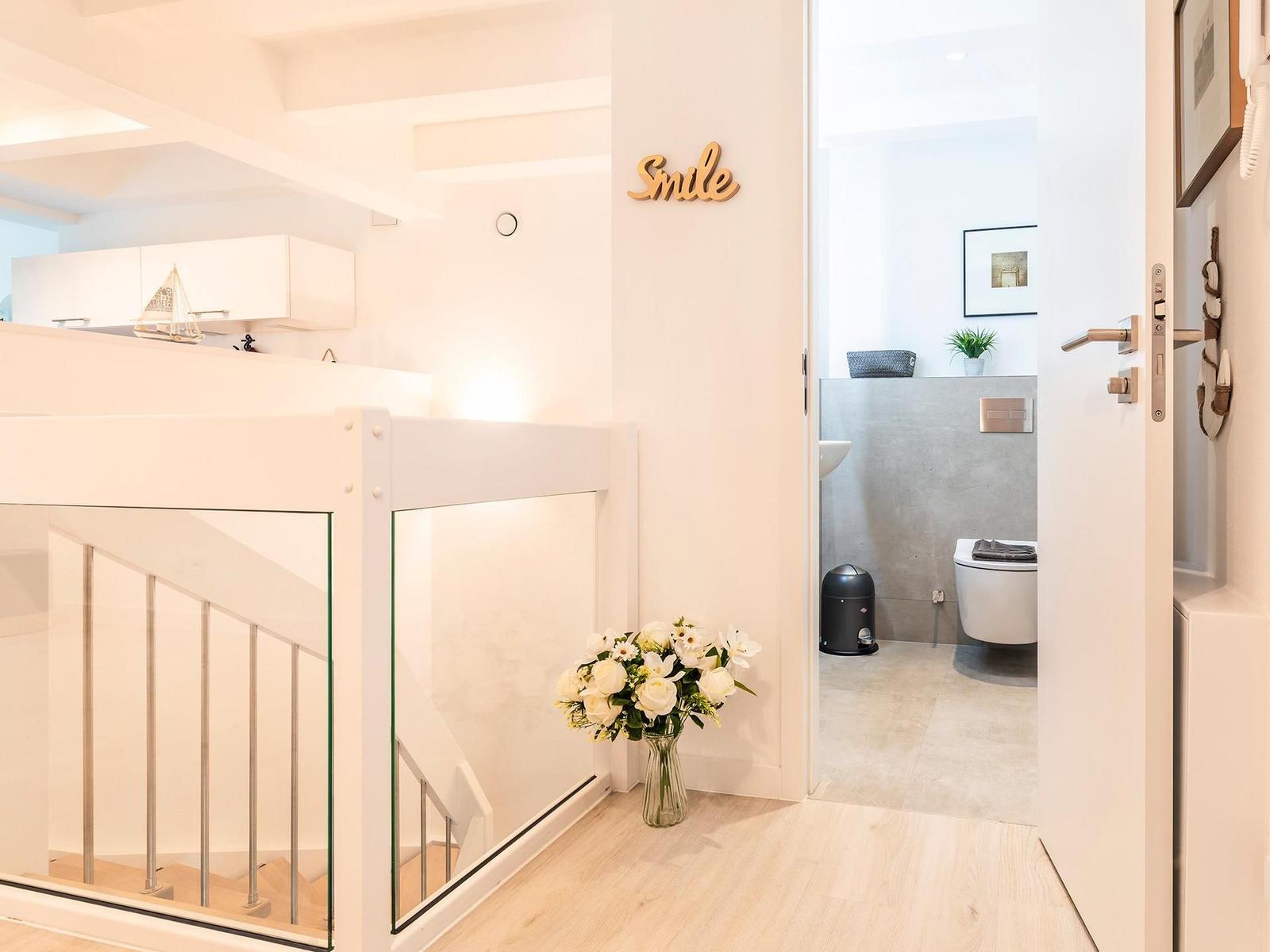 Helle Eingangshalle mit Treppe, Blumen und Blick in die Toilette.