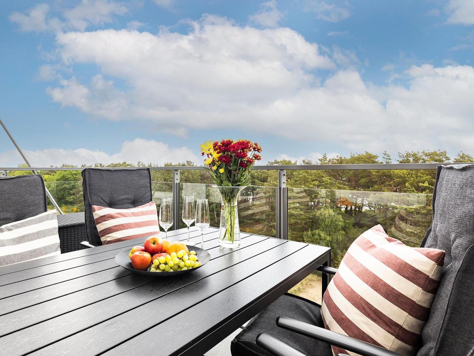 Terrasse mit Tisch, Stühlen und Blumen. Blick auf grüne Bäume und blauen Himmel.
