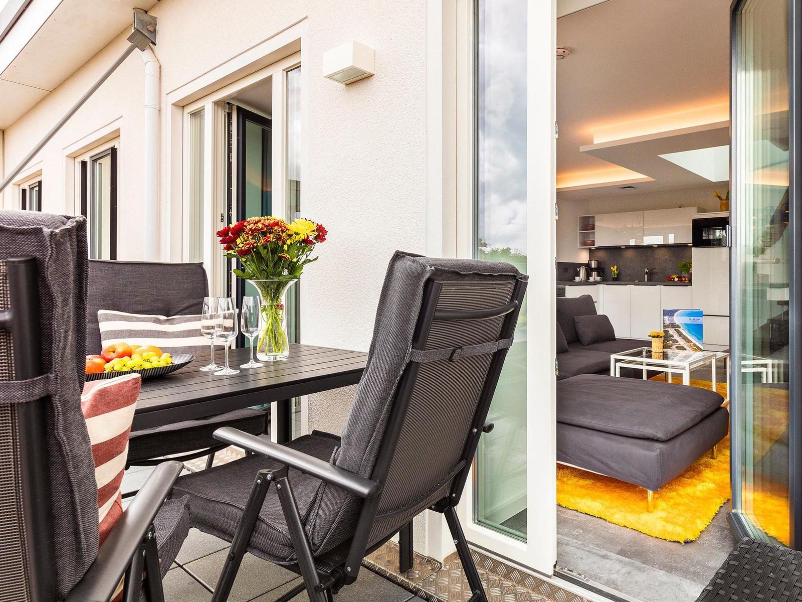 Terrasse mit Tisch, Stühlen und Blumen. Durch Glastüren ist das Wohnzimmer mit Sofa und Küche sichtbar.