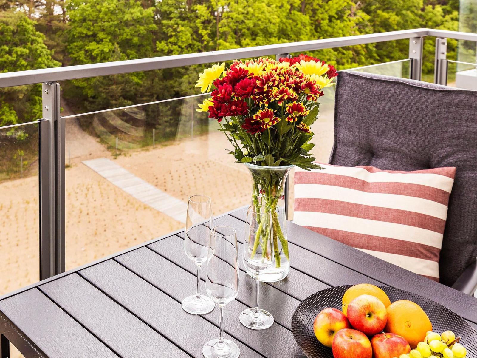 Terrasse mit Tisch, Stühlen, Blumen und Obst. Blick auf Strand und Wald.