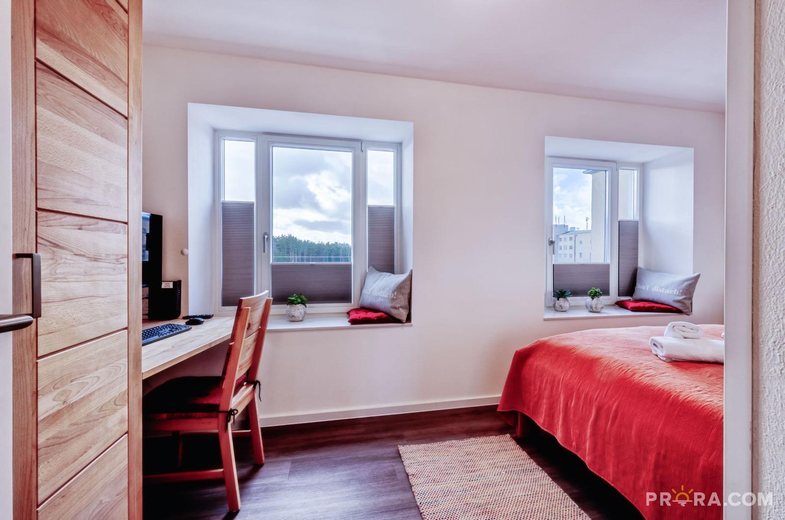 Schlafzimmer mit Bett, Schreibtisch und zwei Fenstern mit Blick auf Außenbereich.