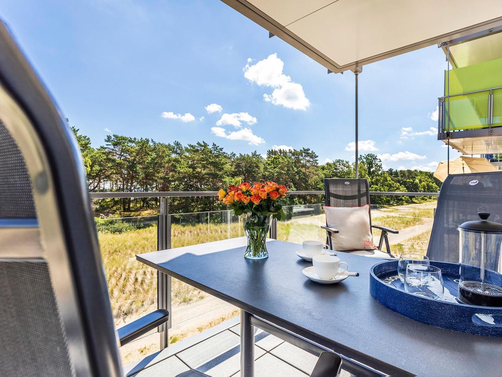 Terrasse mit Tisch, Stühlen und Blick auf Wald. Blumen, Tassen und Kaffeekanne darauf.