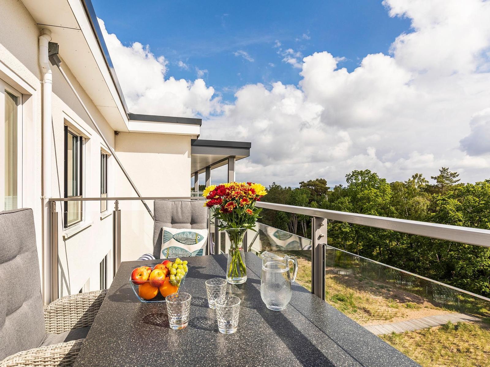 Terrasse mit Tisch, Stühlen, Blumen und Obst. Blick auf Wald und Himmel.