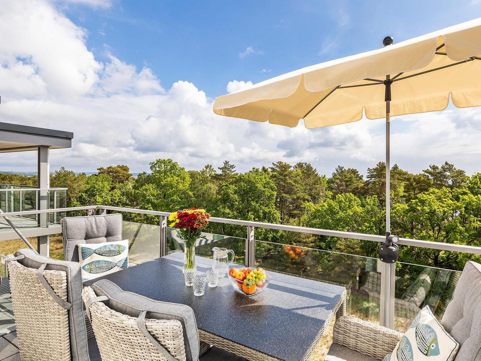 Terrasse mit Tisch, Stühlen und Sonnenschutz. Blick auf Wald und Himmel.