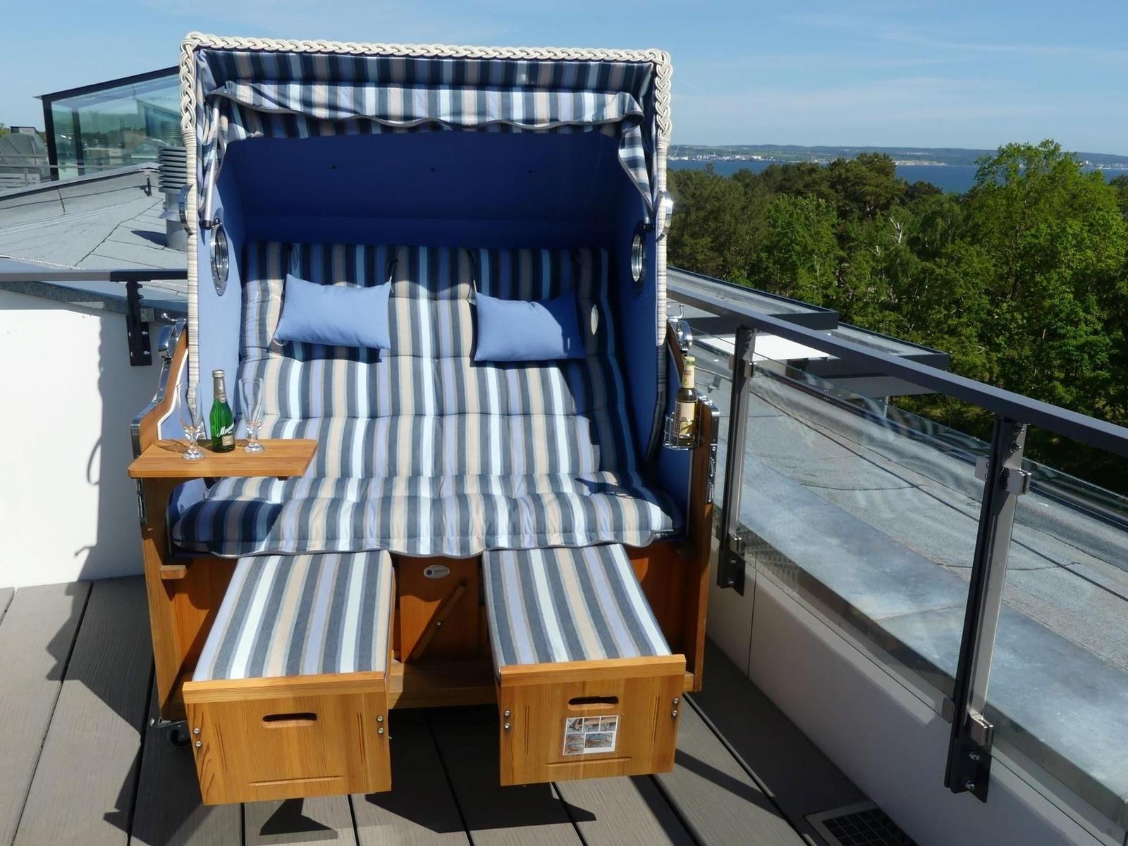Hochwertiger Strandkorb mit blau-weißem Streifenmuster auf Balkon mit Blick auf Wald und Wasser.