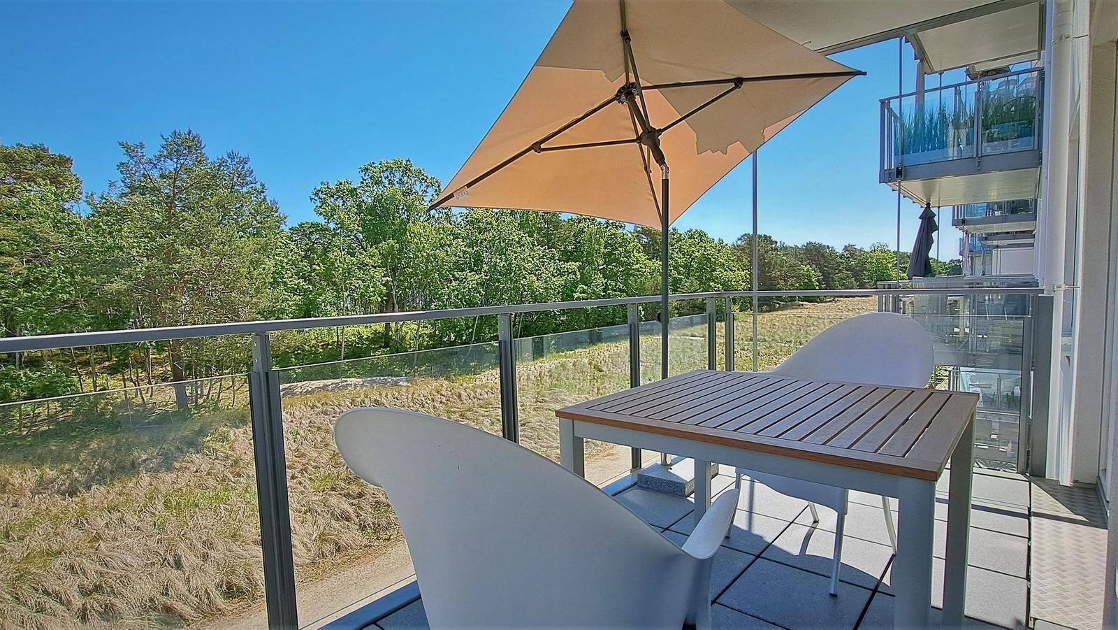 Terrasse mit Tisch, zwei Stühlen und Sonnenschirm. Blick auf Wald und Grasland.