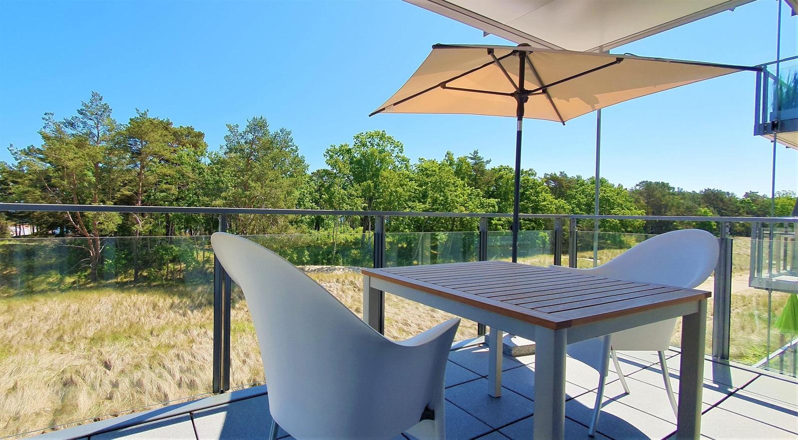 Terrasse mit Tisch, zwei Stühlen und Sonnenschirm. Blick auf Wald und Grasland.