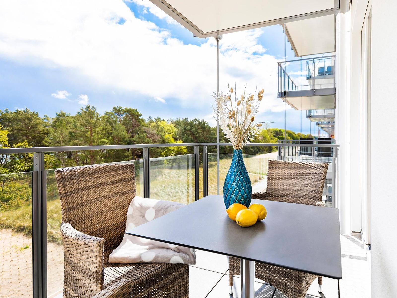 Terrasse mit Tisch, zwei Stühlen und Blumenvase. Blick auf Wald und Himmel.