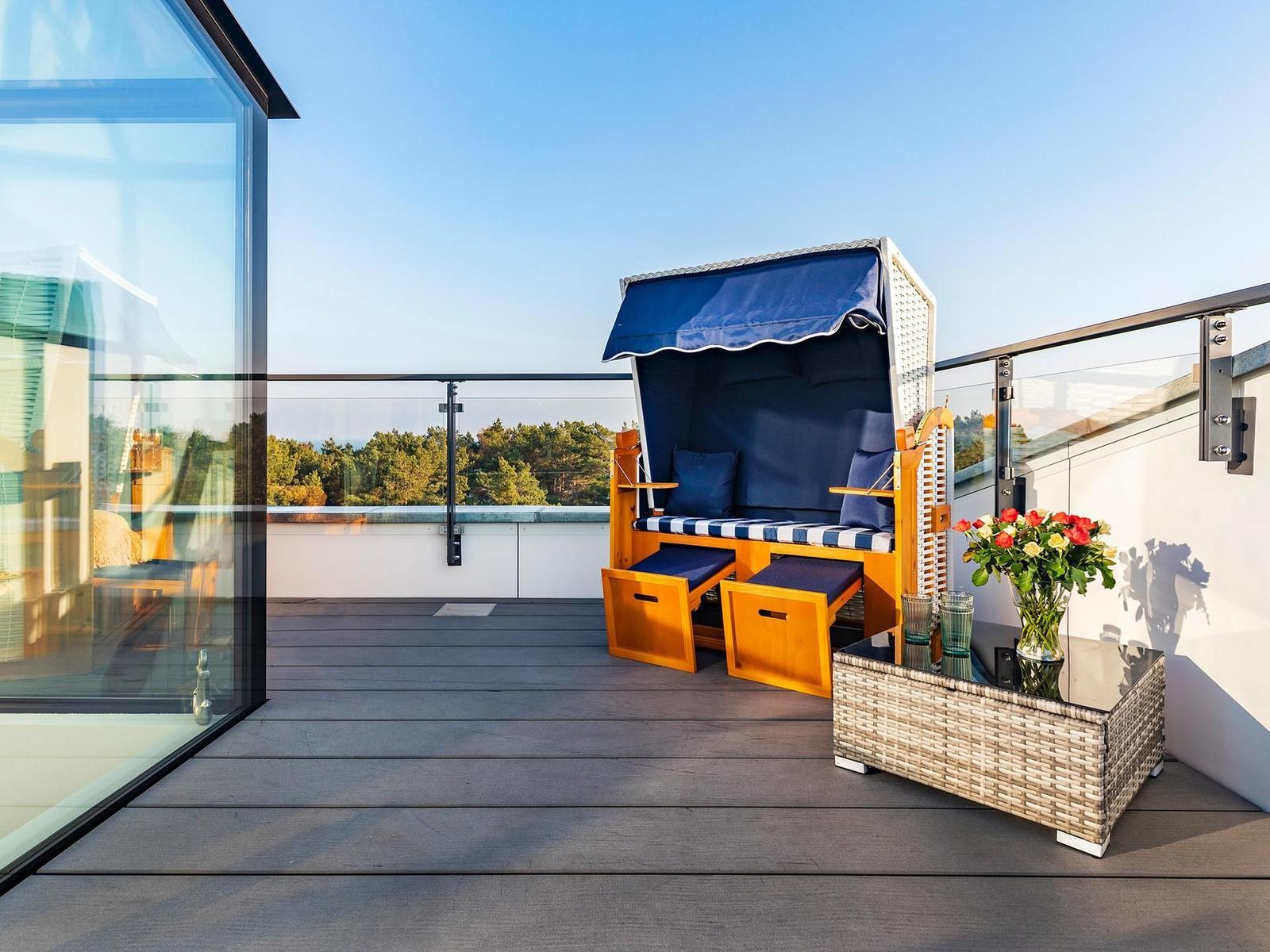 Terrace with beach chair, flowers, and glass railing. View of trees and sky.