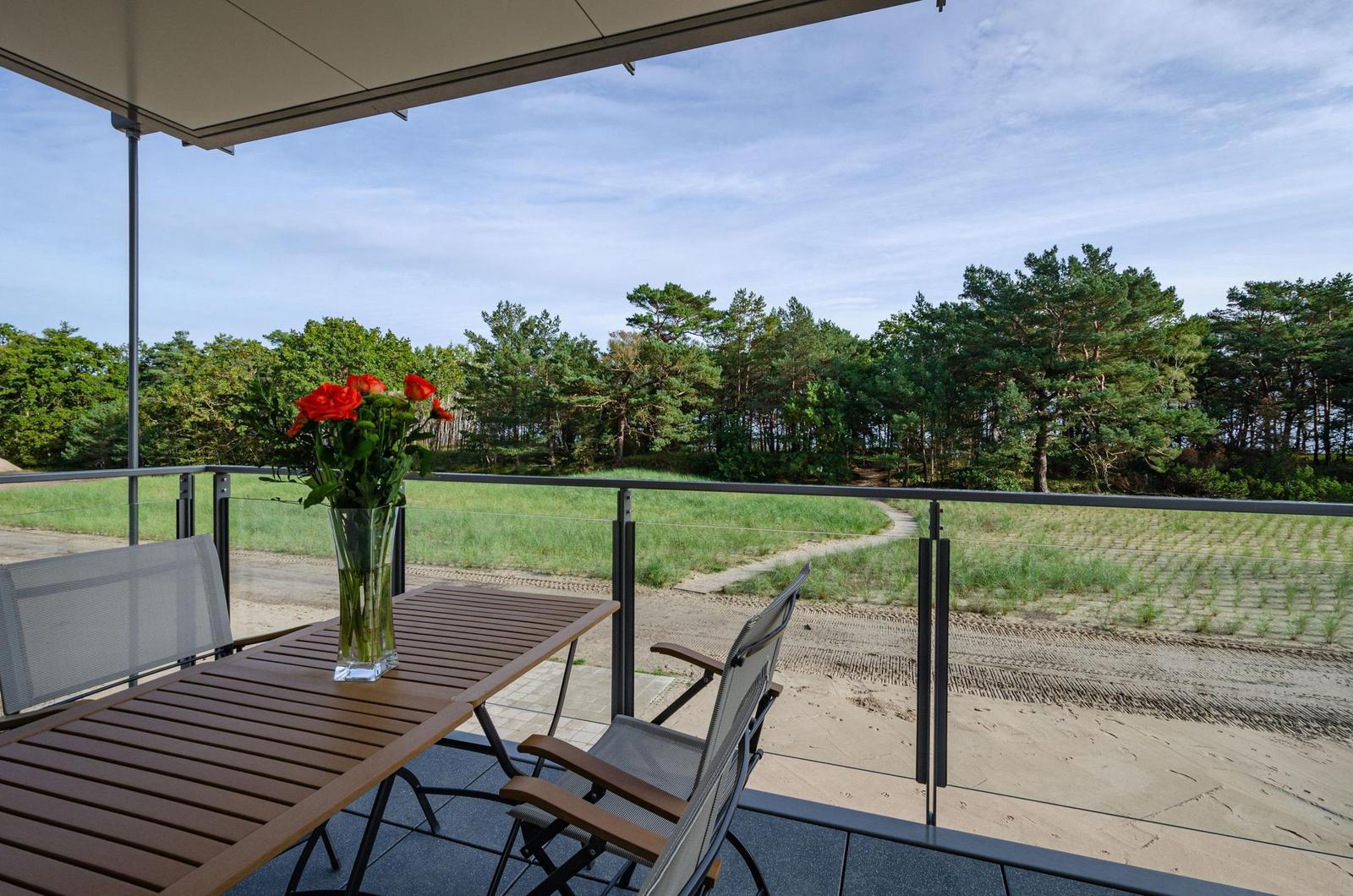 Terrasse mit Tisch, Stühlen und Blumen. Blick auf Wald und Sandstrand.