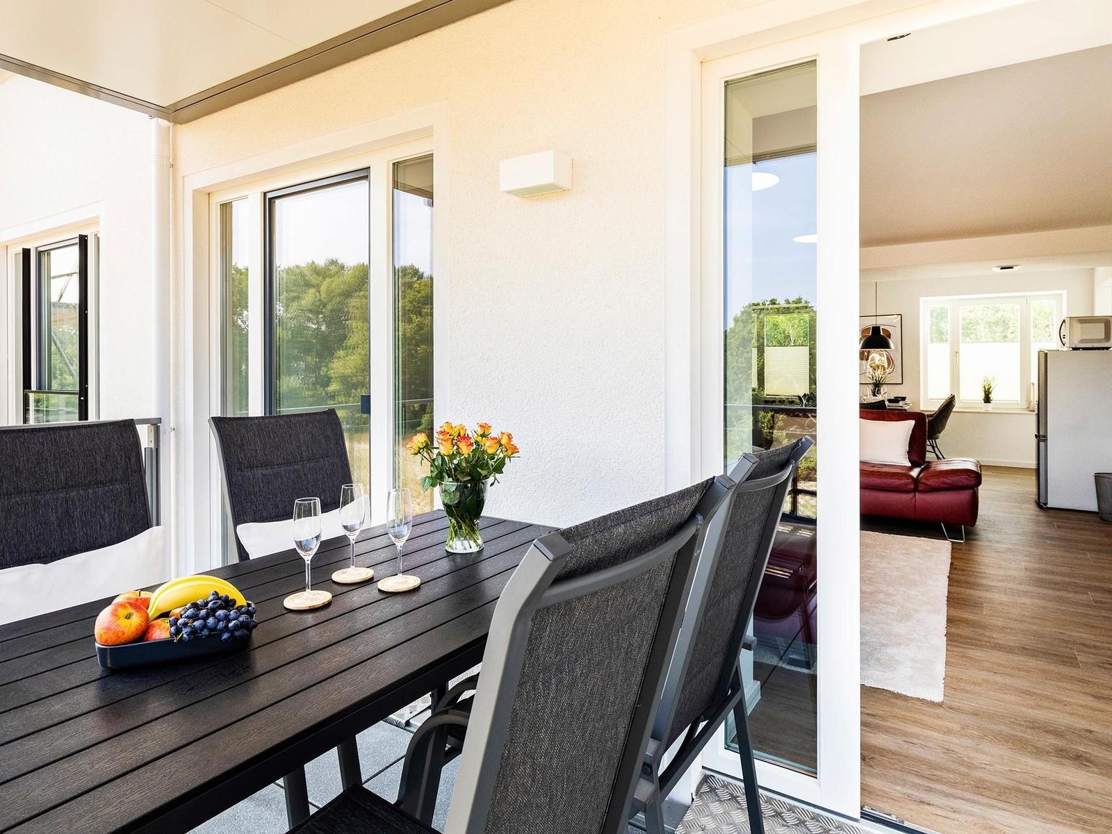 Terrasse mit Tisch, Stühlen und Obst. Durchgang ins Wohnzimmer mit rotem Sofa.