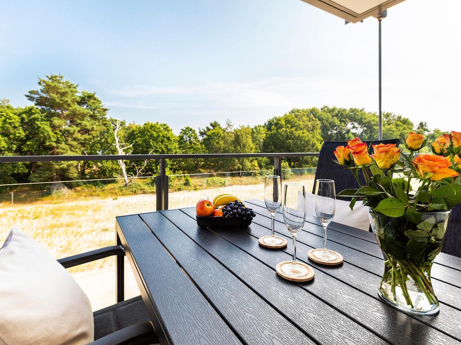 Terrasse mit Tisch, Stühlen, Blumen und Obst. Blick auf Wald und Feld.