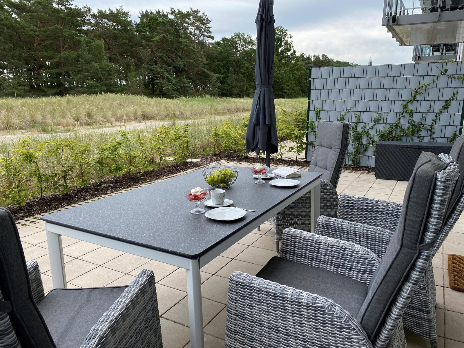 Terrasse mit Tisch, Stühlen und Obst. Hintergrund: Grünfläche und Wald.