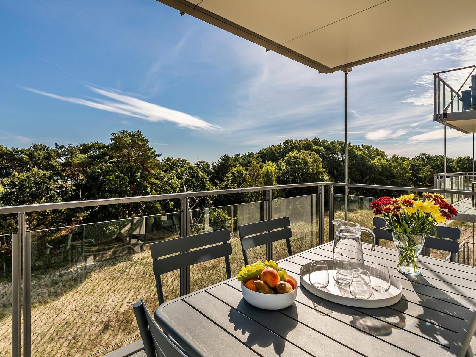 Terrasse mit Tisch, Stühlen, Obst und Blumen. Blick auf Wald unter blauem Himmel.