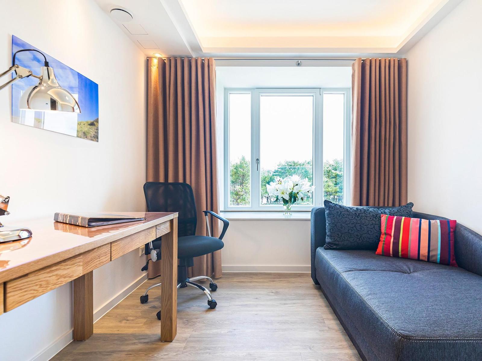 Ein Raum mit Schreibtisch, Stuhl, Sofa und Fenster mit Blick auf Bäume.