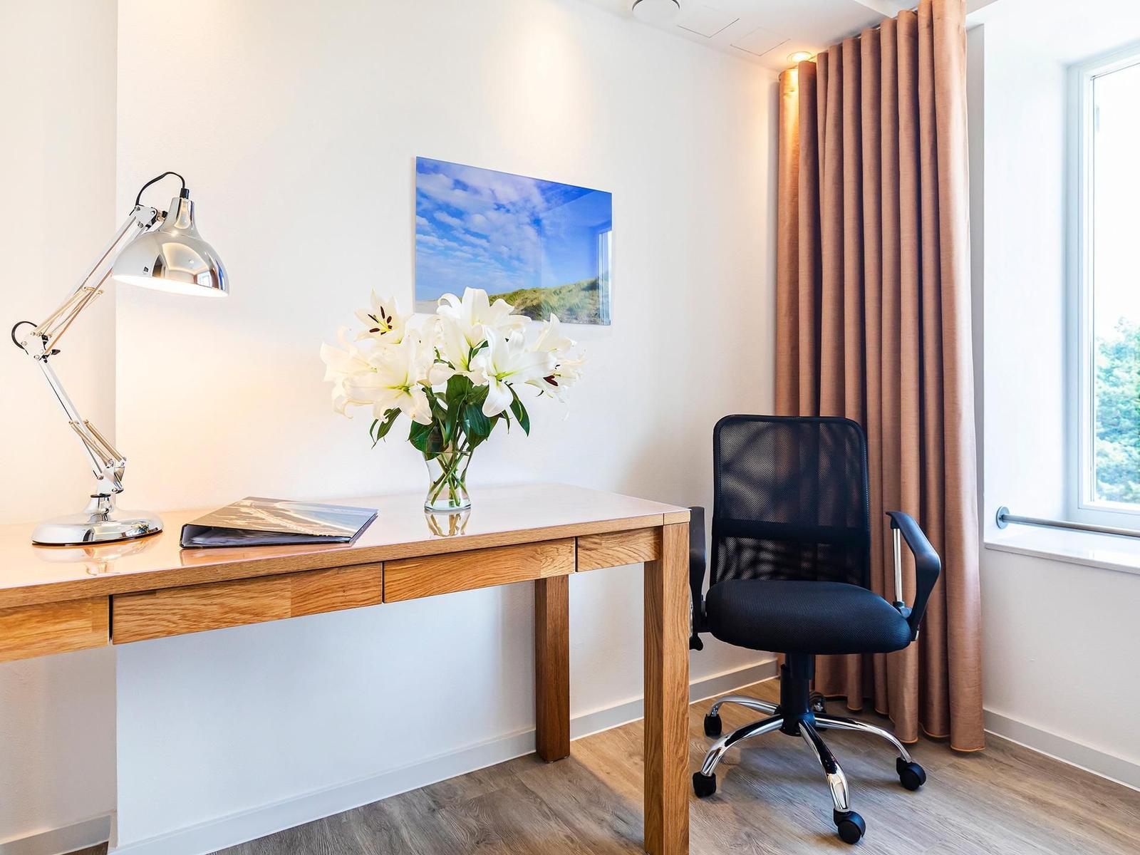 Schreibtisch mit Stuhl, Blumen und Lampe vor Fenster mit Vorhang.