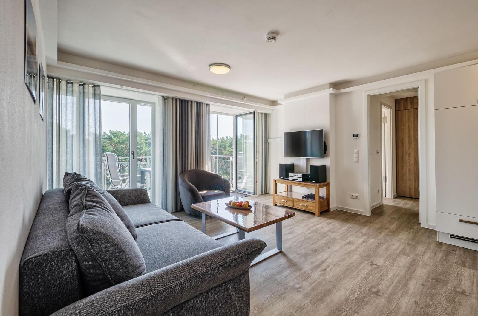 Wohnzimmer mit Sofa, Couchtisch und TV. Balkon mit Sicht auf Bäume.