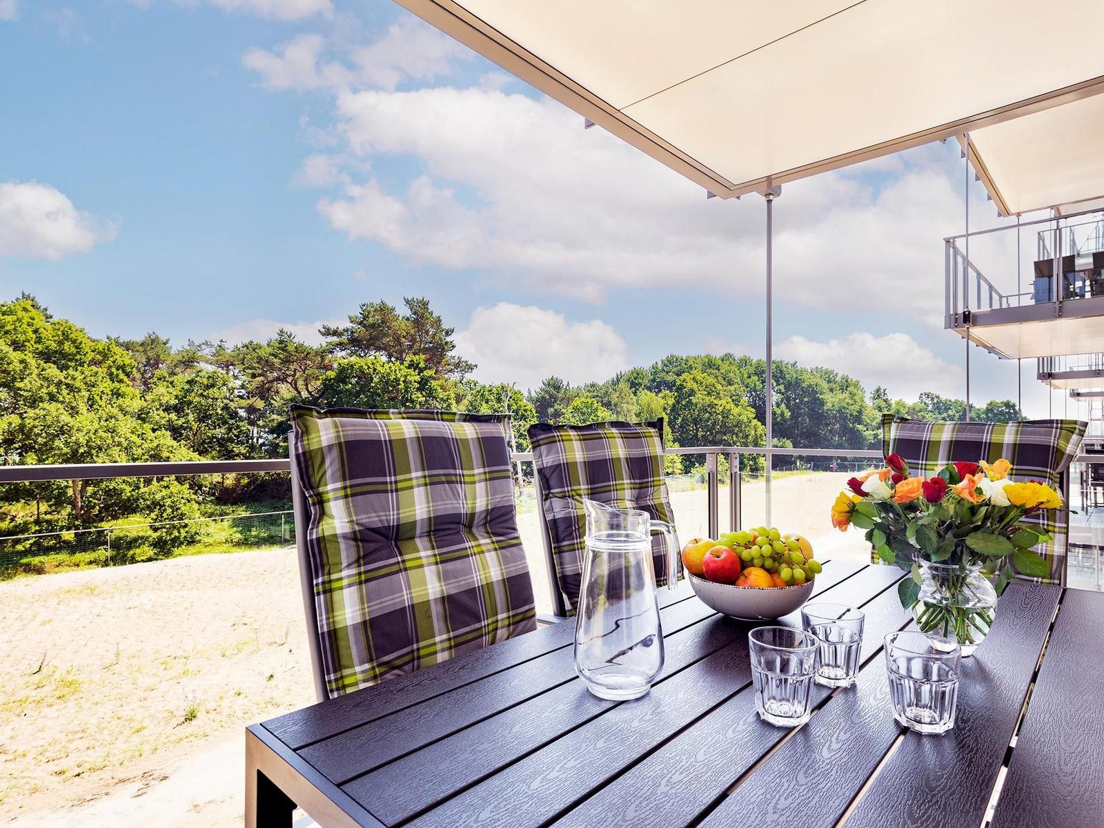 Terrasse mit Tisch, Stühlen und Obst. Blick auf Wald und Himmel.