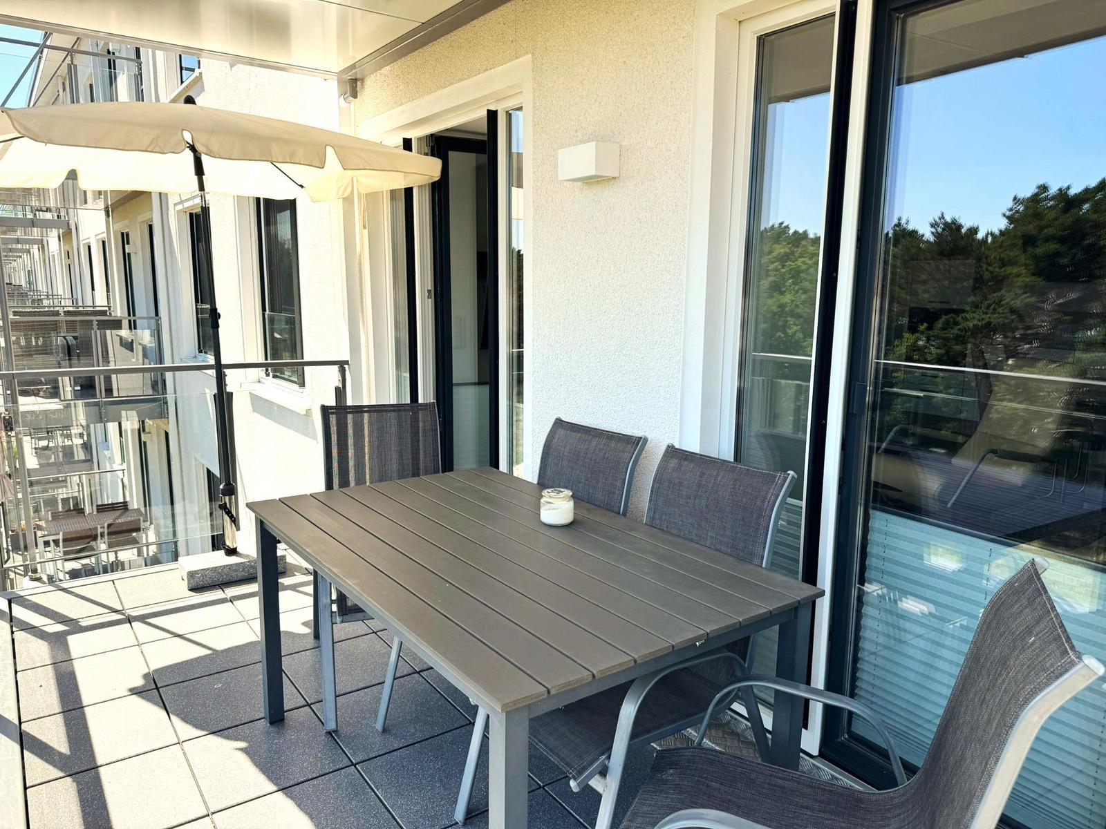 Terrasse mit Tisch, Stühlen und Sonnenschirm. Glasgeländer und Schiebetüren zum Innenraum.