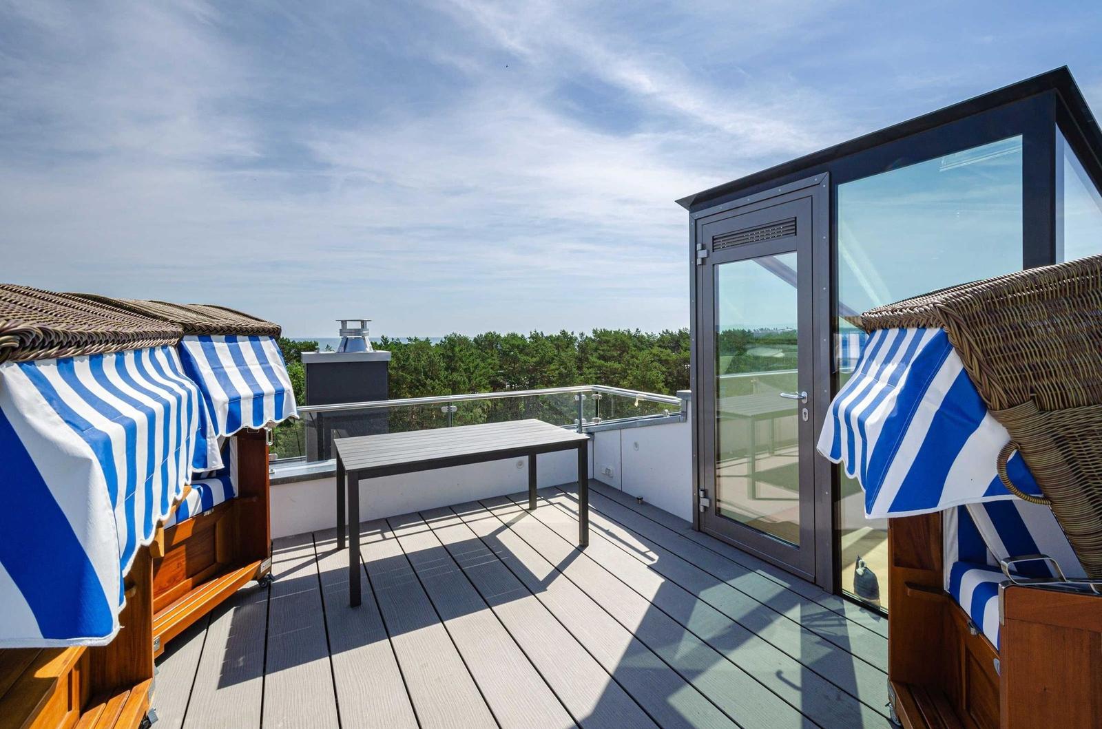 Terrasse mit Strandkörben, Tisch und Blick auf Wald und Meer.