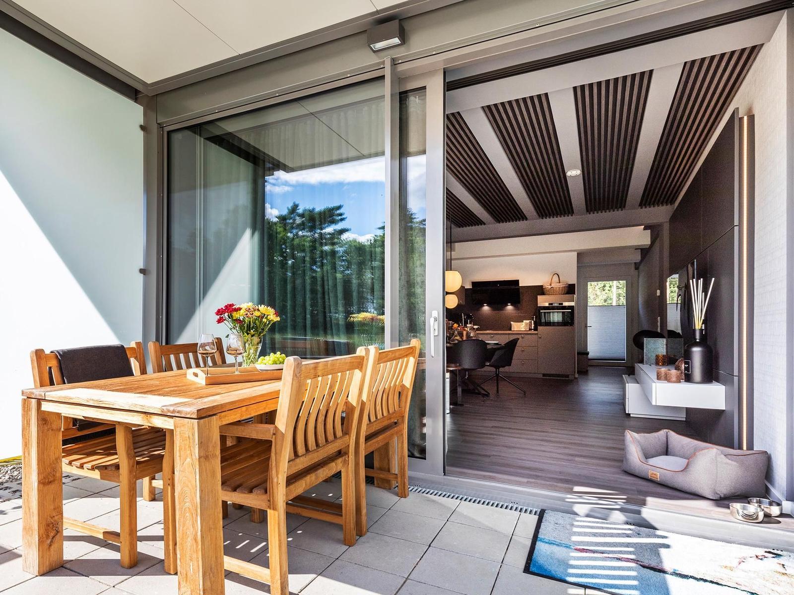 Terrasse mit Holztisch und Stühlen. Durch Glastüren ist Küche und Wohnbereich sichtbar.