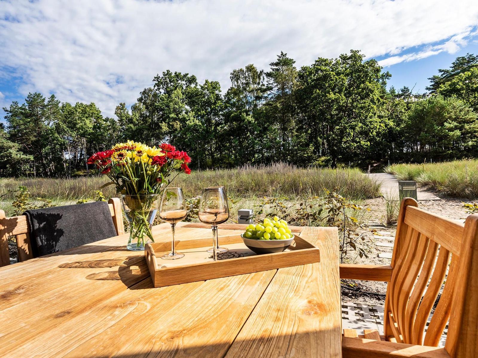 Holztisch mit Weinflaschen und Obst im Freien, Blick auf Wald und Strand.
