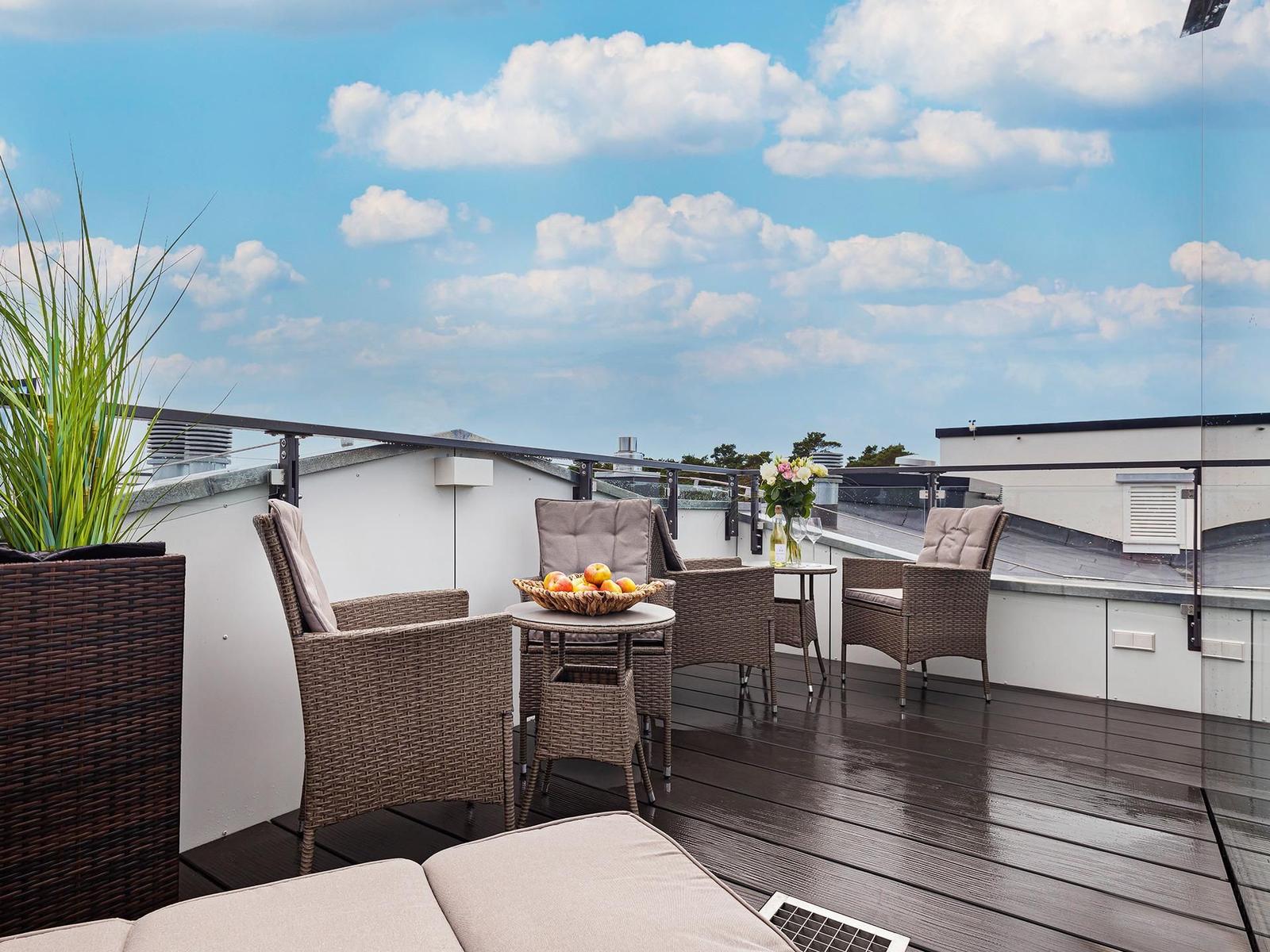 Terrasse mit Sitzgelegenheiten, Tisch und Pflanze unter blauem Himmel mit Wolken.