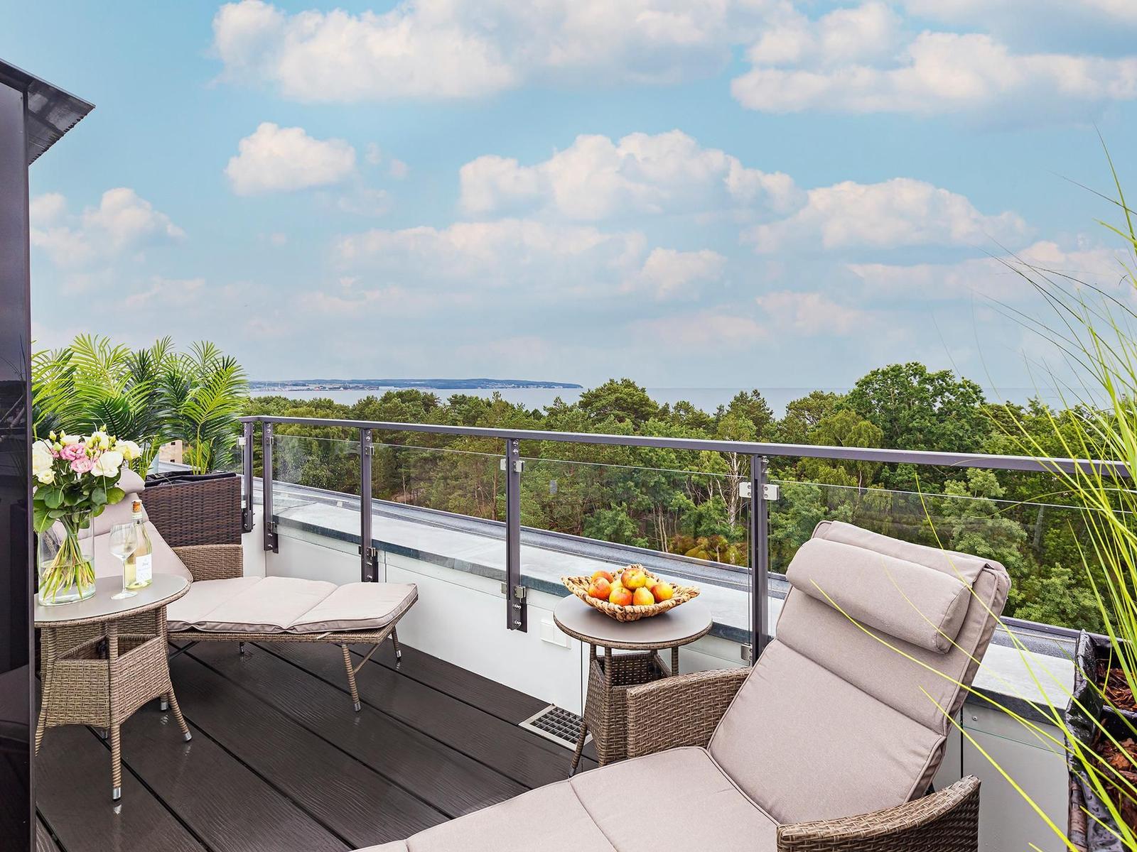 Terrasse mit Liege, Tisch und Obstschale. Blick auf Wald und See unter blauem Himmel.