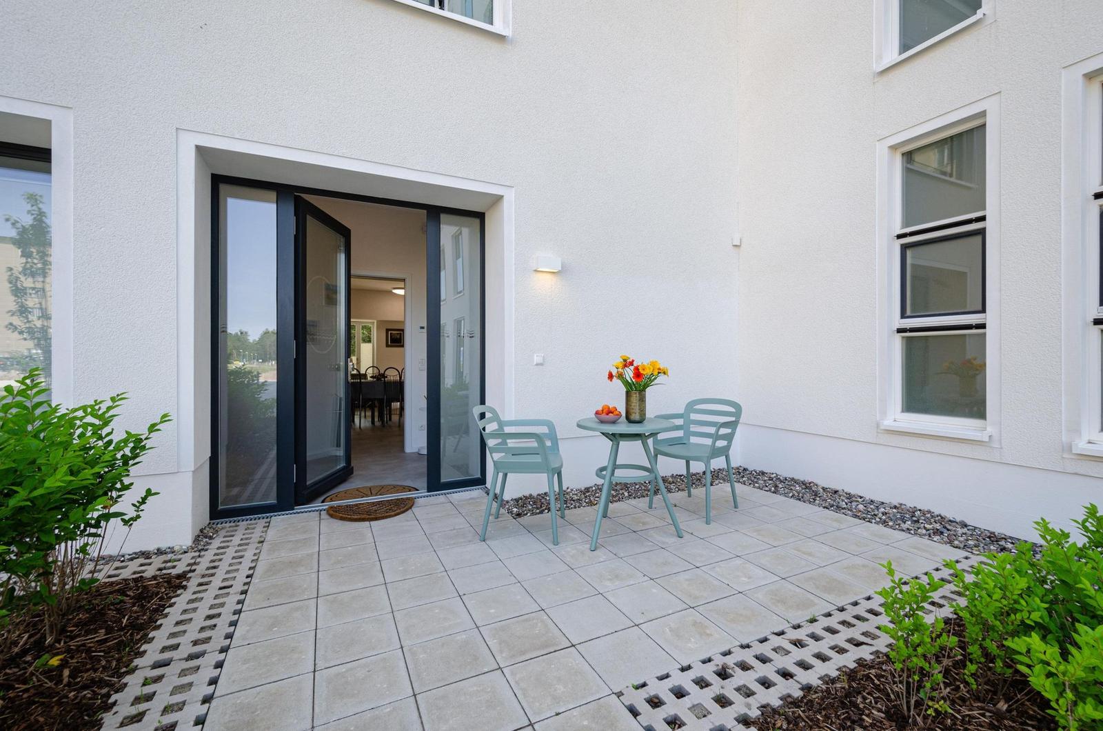 Terrasse mit Tisch, zwei Stühlen und Blumen. Durchgehendes Glas führt ins Haus.