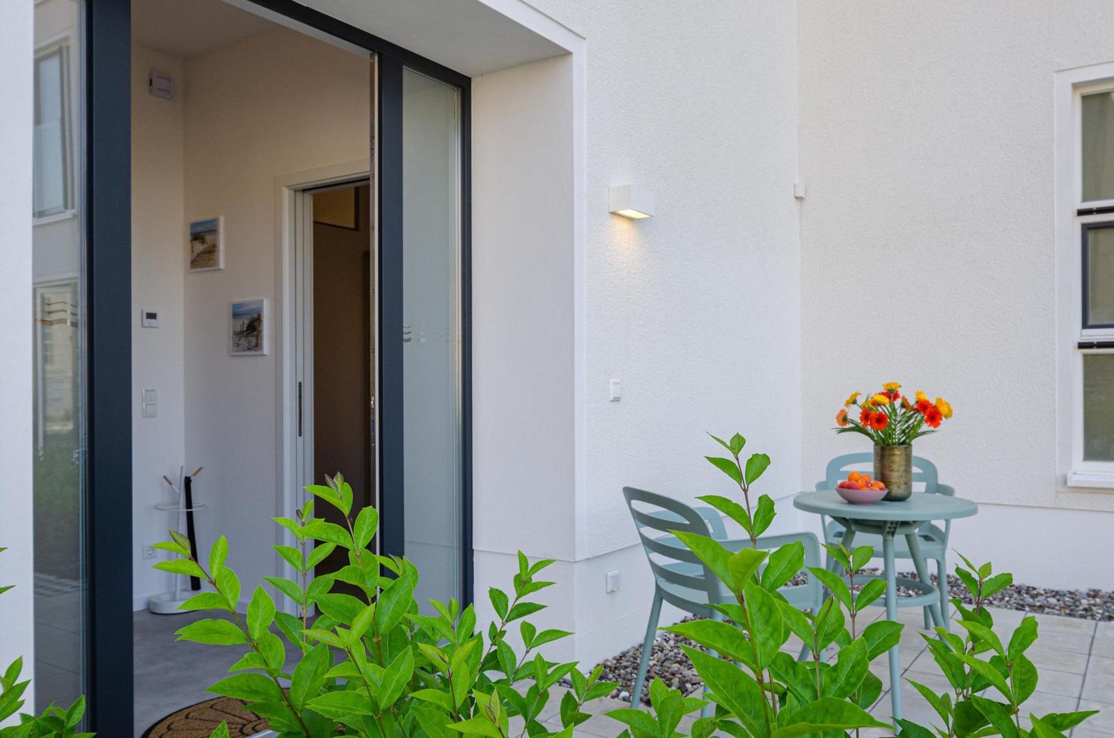 Terrasse mit Tisch, Stühlen und Blumen. Durchgehendes Glas führt ins Haus.